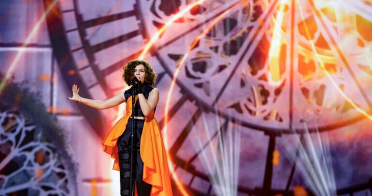 A woman in an orange dress singing in front of a clock during her performance.