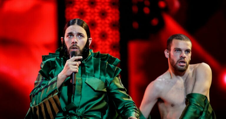 Two men in green costumes singing into a microphone with vibrant energy and harmonious voices.